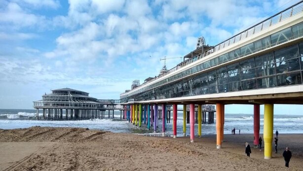 A pier with colorful columns extending into the sea, a beach in the foreground, and a large building with a glass facade in the background. - Ground Floor Apartment near Scheveningen (The Hague)