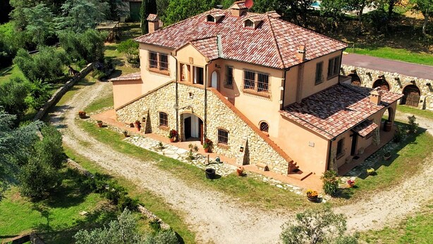 Un grand bâtiment en pierre à deux étages, surmonté d’un toit en tuiles, entouré d’un chemin de gravier et d’une végétation luxuriante.