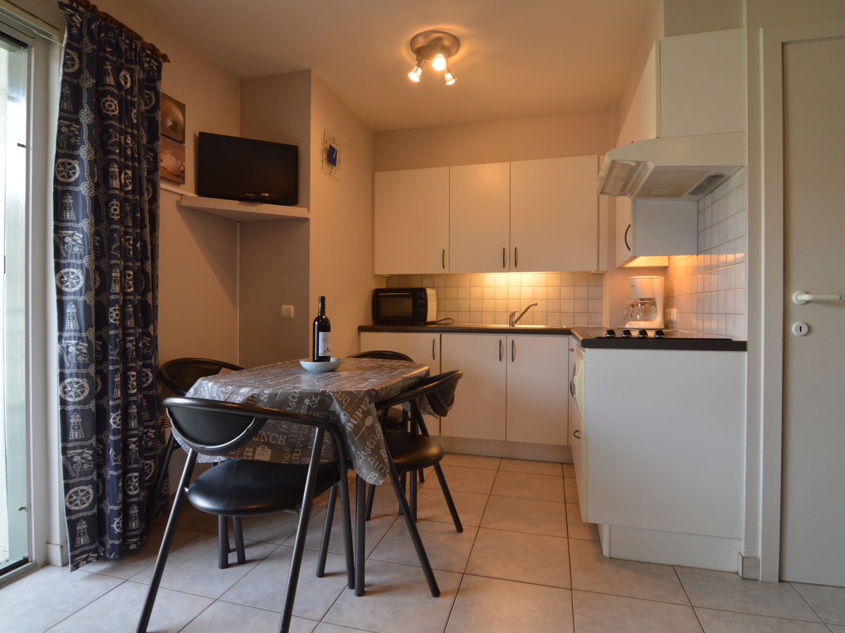 Une cuisine compacte avec des meubles blancs, un four à micro-ondes, un évier et un réfrigérateur. On y trouve une table à manger avec des chaises et une bouteille de vin posée dessus.