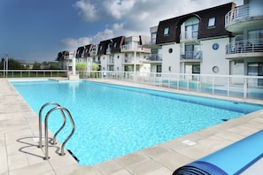 A swimming pool with a metal ladder, surrounded by a white fence, and adjacent to a multi-story building with balconies.