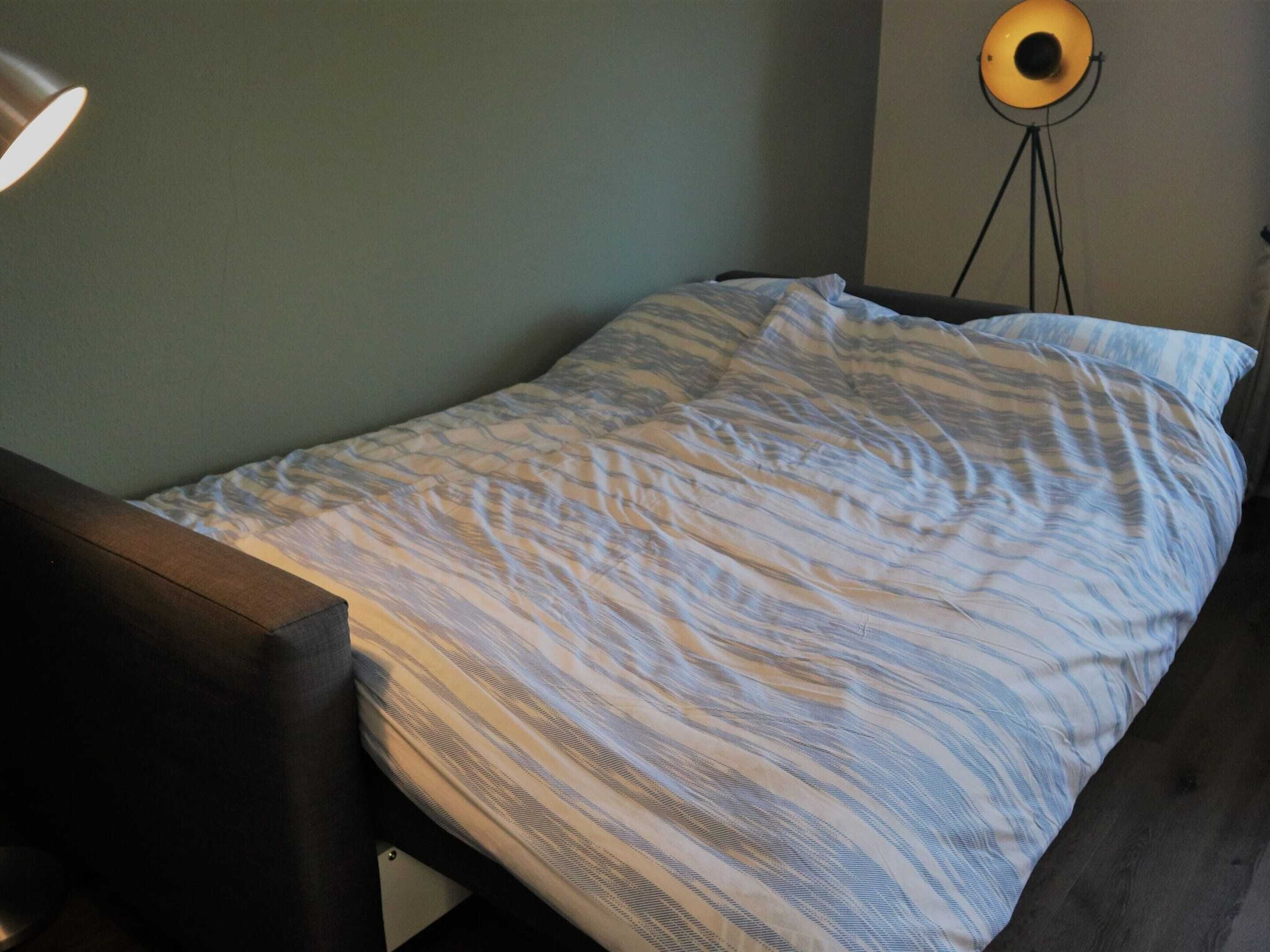 Una cama individual con una colcha a rayas en una habitación con pared verde y una lámpara de pie.