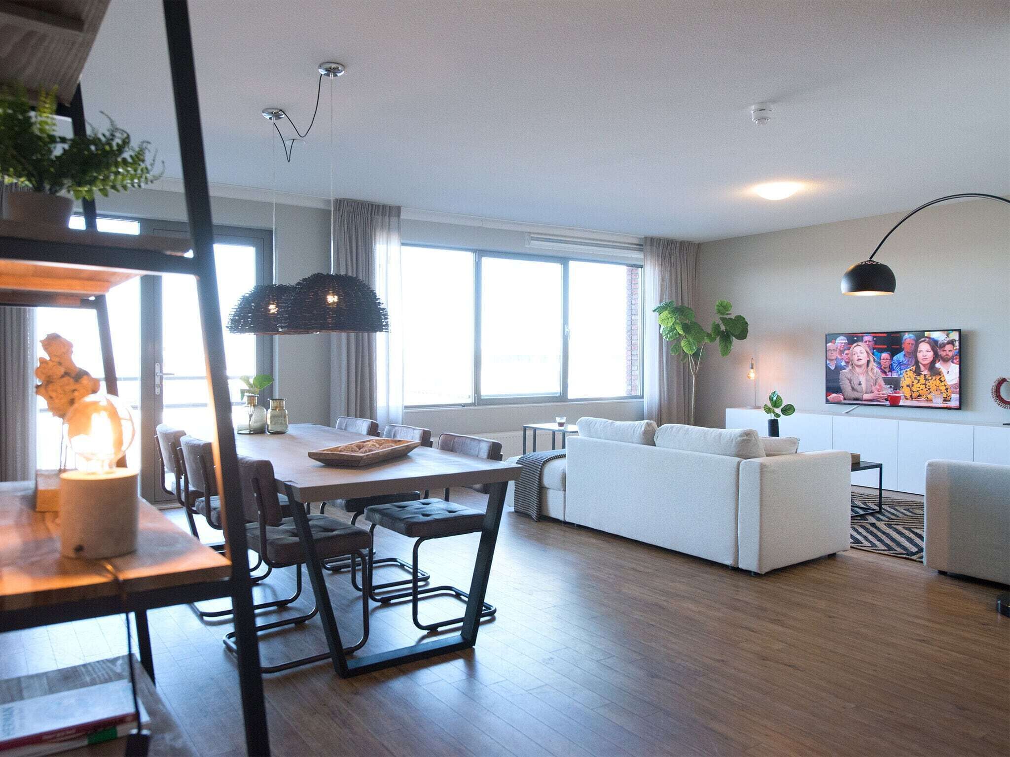 A modern living room with a sofa, dining table, and chairs. There is a TV mounted on the wall and a window providing natural light.