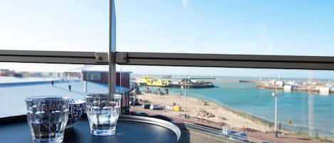 En balkong med ett bord och två glas vatten, med utsikt över en strand och en hamn med fartyg och kranar.