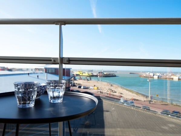 A balcony with a table and two glasses of water, overlooking a beach and port with ships and cranes. - Apartment in Den Haag for 4 Person (The Hague)