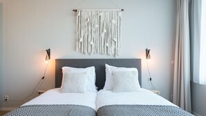 A double bed with a grey headboard, two bedside tables with lamps, a macrame wall hanging, and a window with curtains. - Apartment in Den Haag for 4 Person (The Hague)