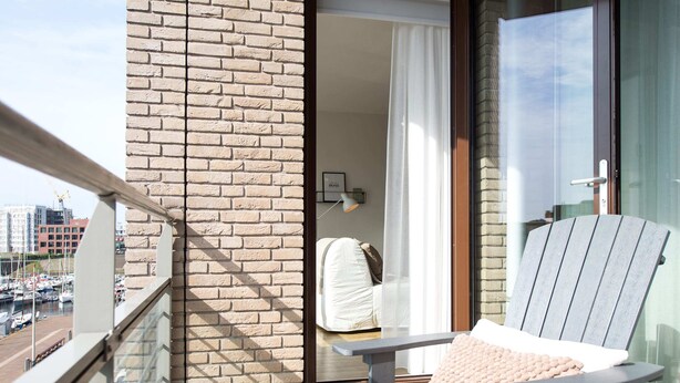 A balcony with a view of a marina, a brick wall, and a sliding glass door leading to a room with a bed and a lamp. - Apartment in Netherlands Near the Beach (The Hague)