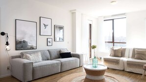 A modern living room with a grey sofa, a round wicker coffee table, and a large window offering a city view. - Apartment in Netherlands Near the Beach (The Hague)