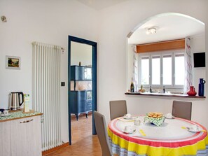 A dining area with a round table covered in a striped tablecloth, chairs, a window with curtains, a radiator, a cabinet with glass doors, and a kitchen area with a kettle and a coffee machine. - Apartment in Casale With Panoramic Vineyard Views (Moncucco Torinese)