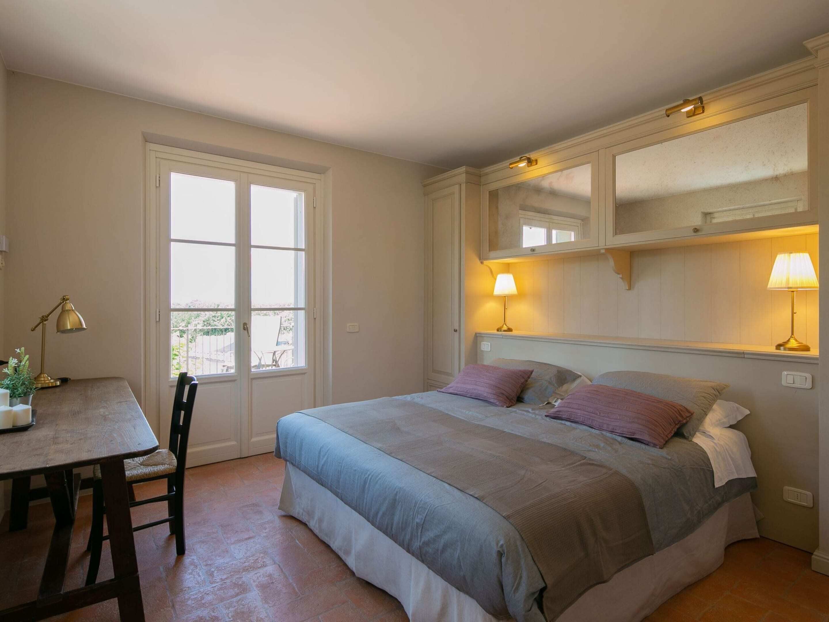 A bedroom with a bed, bedside lamps, a window, a door, and a small table with a lamp.