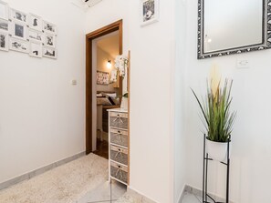 A hallway with a white wall, a wooden door, a white floor, a white cabinet with baskets, a mirror, and a potted plant. - Modern Holiday Home in Zadar With Terrace (Zadar)