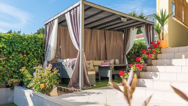 A covered outdoor seating area with a sofa and table, surrounded by potted plants and a small garden. - Modern Holiday Home in Zadar With Terrace (Zadar)