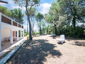 A covered outdoor seating area with tables and chairs, surrounded by trees and a gravel path.