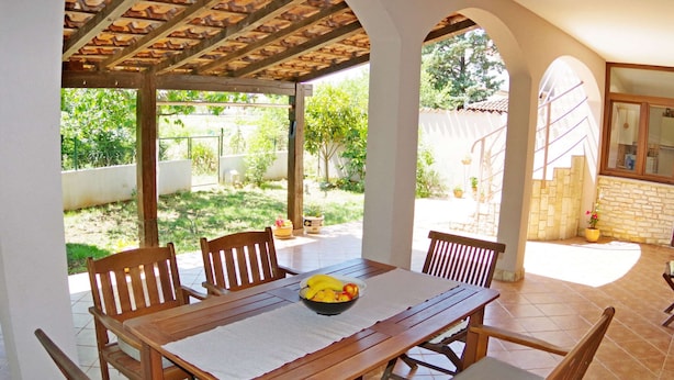 A covered patio with a wooden table and chairs, a bowl of fruit, and a view of the garden. - Apartment in Valbandon With Terrace (Fazana)