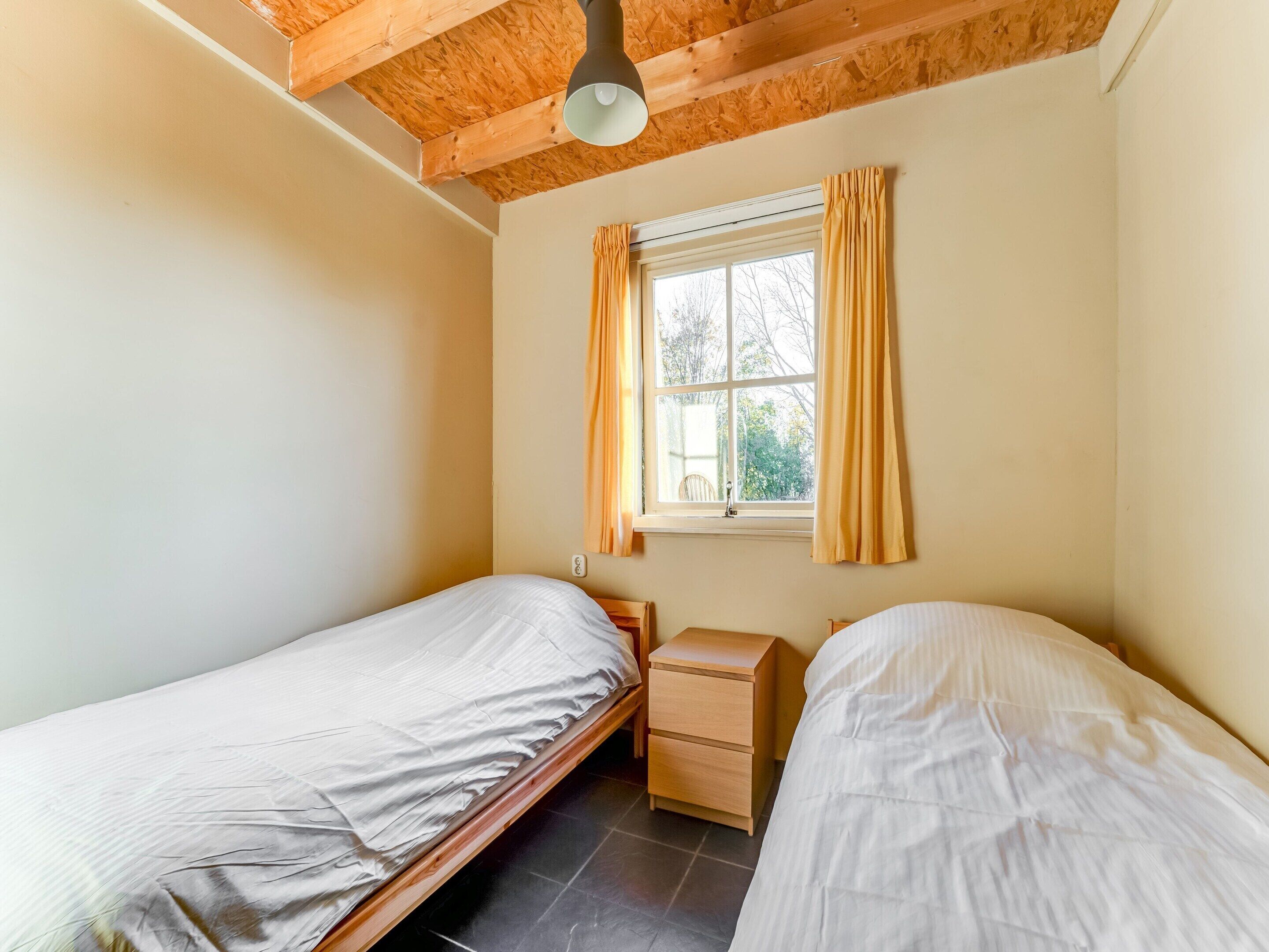 A small, single-room bedroom with two single beds, a wooden nightstand, a window with curtains, and a ceiling fan.