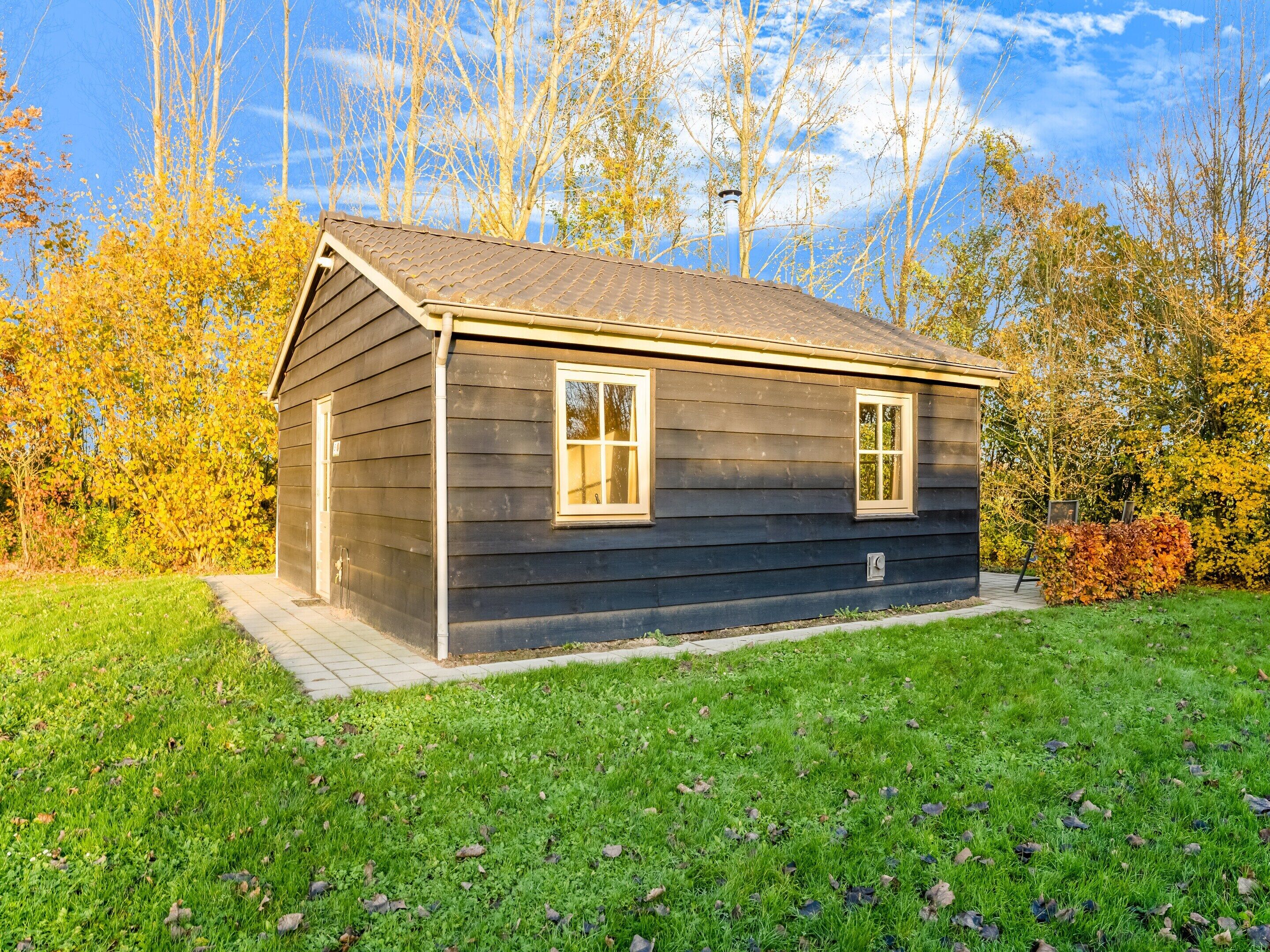 A small, dark wooden cabin with two windows and a sloped roof, surrounded by a grassy yard and autumn-colored trees.
