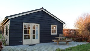 Een donker houten huis met een betegeld terras en twee ramen.