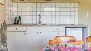 Een kleine keuken met witte kastjes, een wastafel en een kleine koelkast. Er staat een houten stoel bij een tafel met een tafelkleed met bloemen.