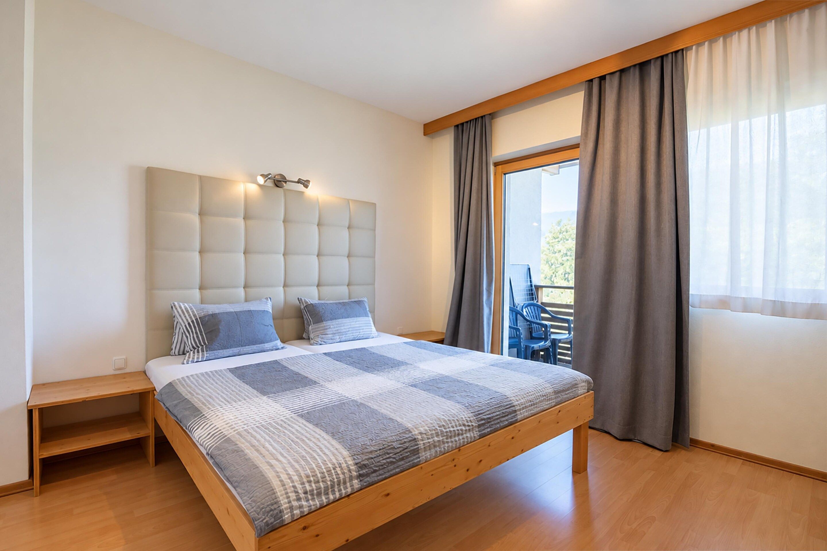 A bedroom with a bed, bedside tables, a headboard with lights, a window with curtains, and a view of the outdoors.