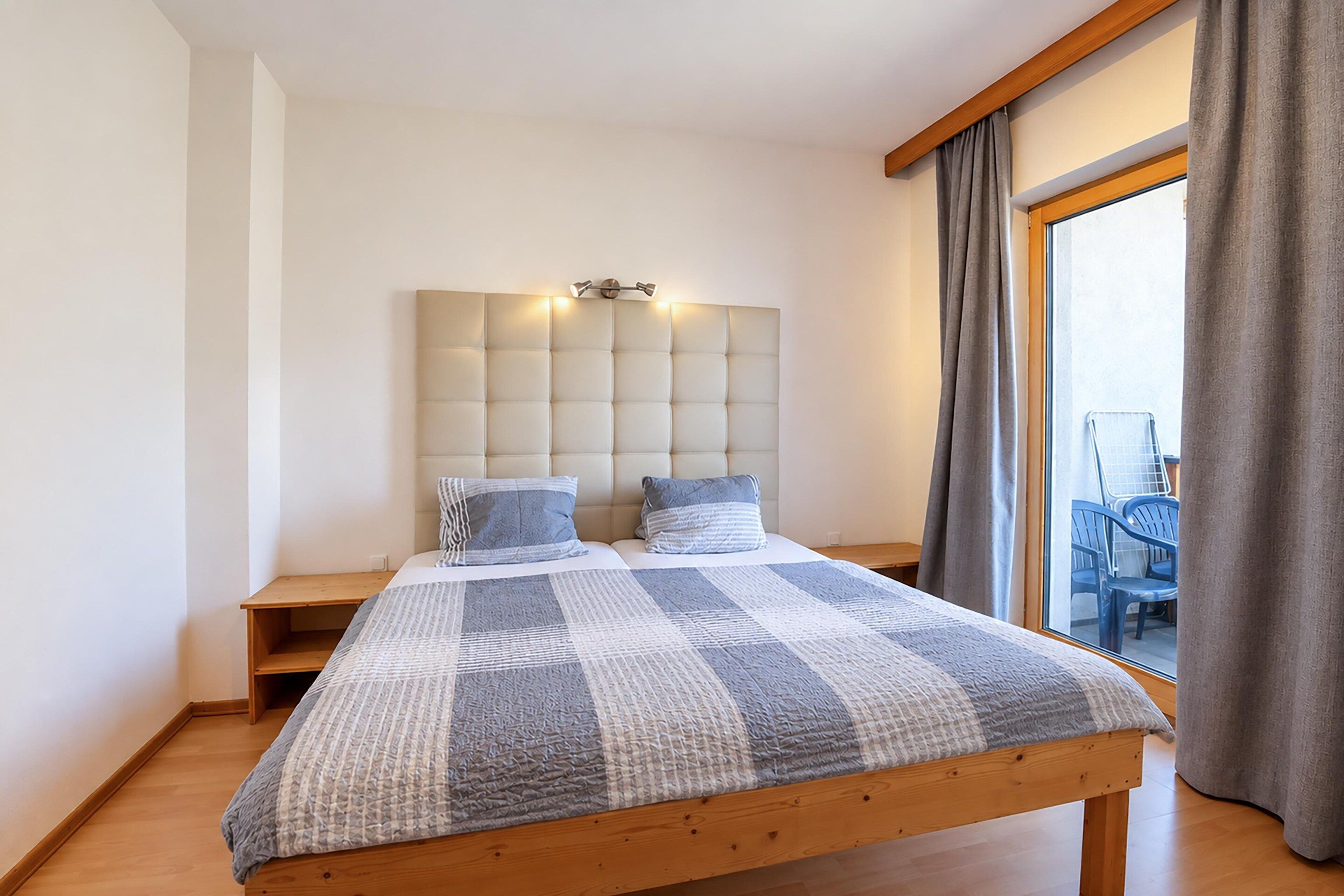 Une chambre à coucher avec un grand lit, une tête de lit dotée de lampes, une table de chevet en bois et une vue sur l’extérieur par une fenêtre ornée de rideaux.