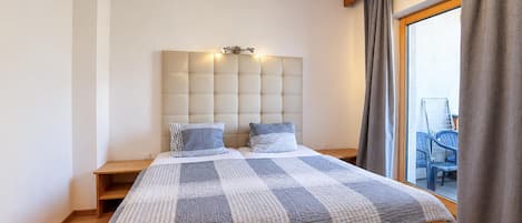 A bedroom with a large bed, a headboard with lights, a wooden nightstand, and a view of the outside through a window with curtains.