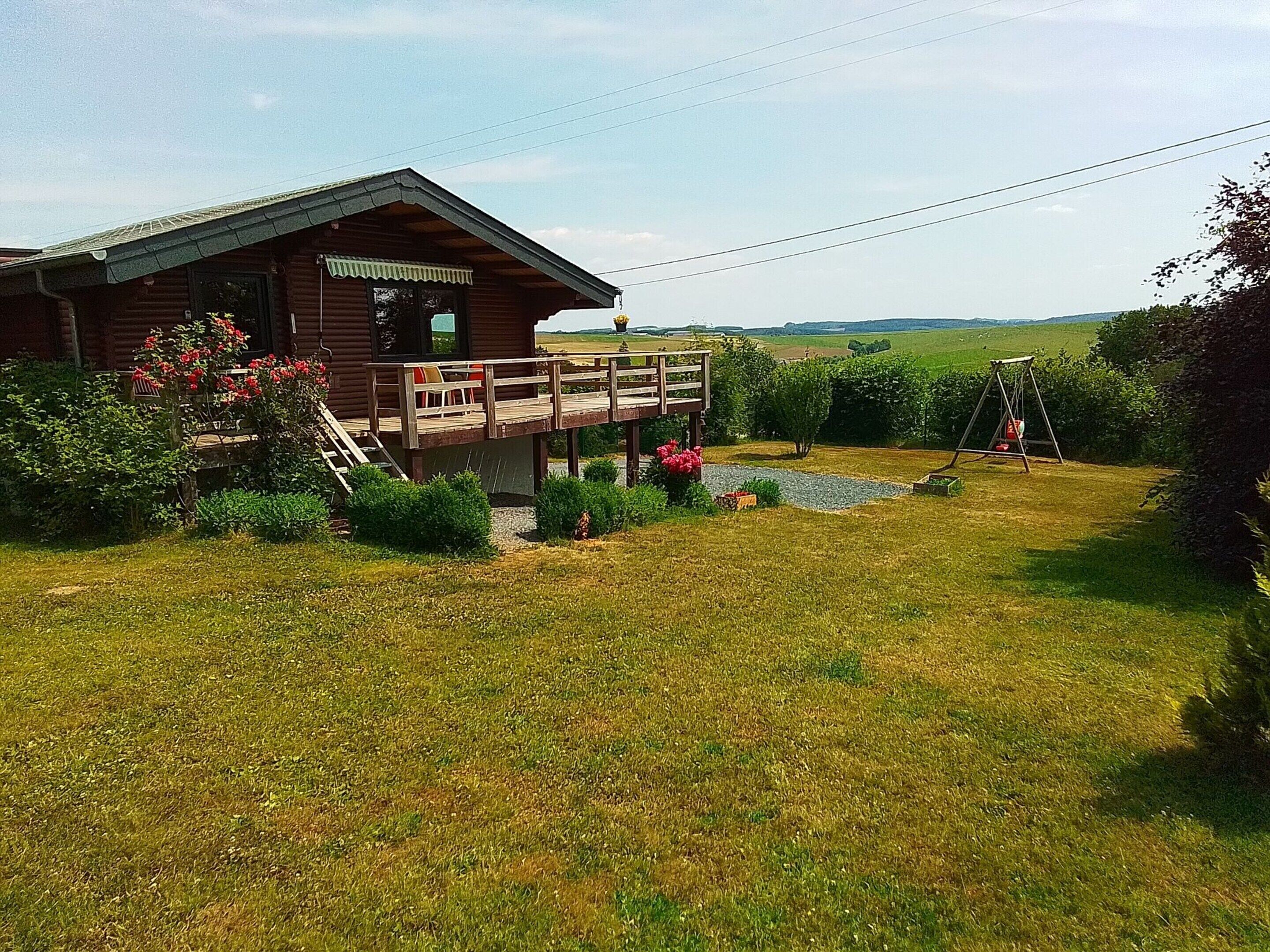 A wooden cabin with a deck, surrounded by a grassy yard and a swing set.