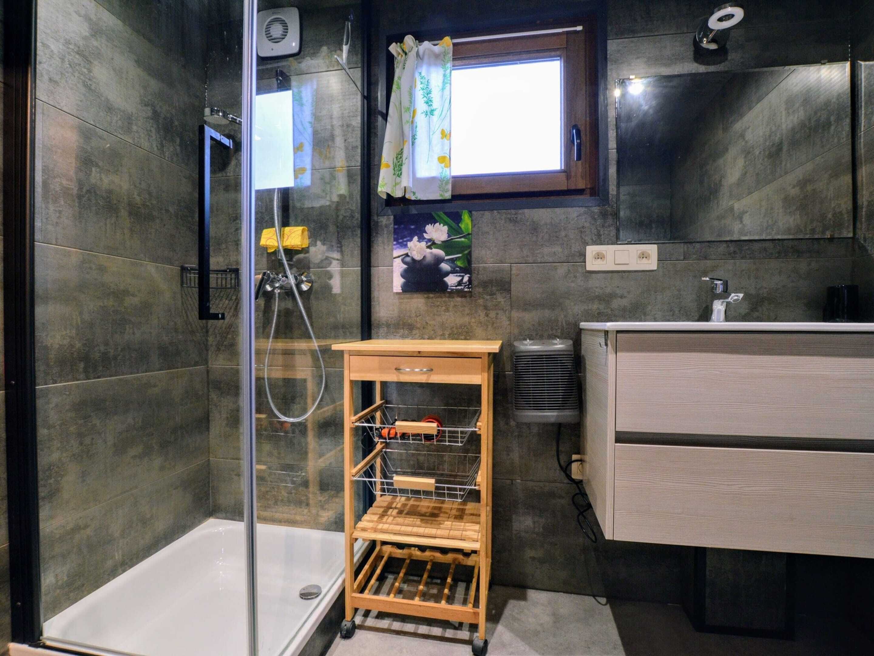 A modern bathroom with a glass-enclosed shower, a wooden drying rack, a small window with a curtain, and a sink with a mirror.