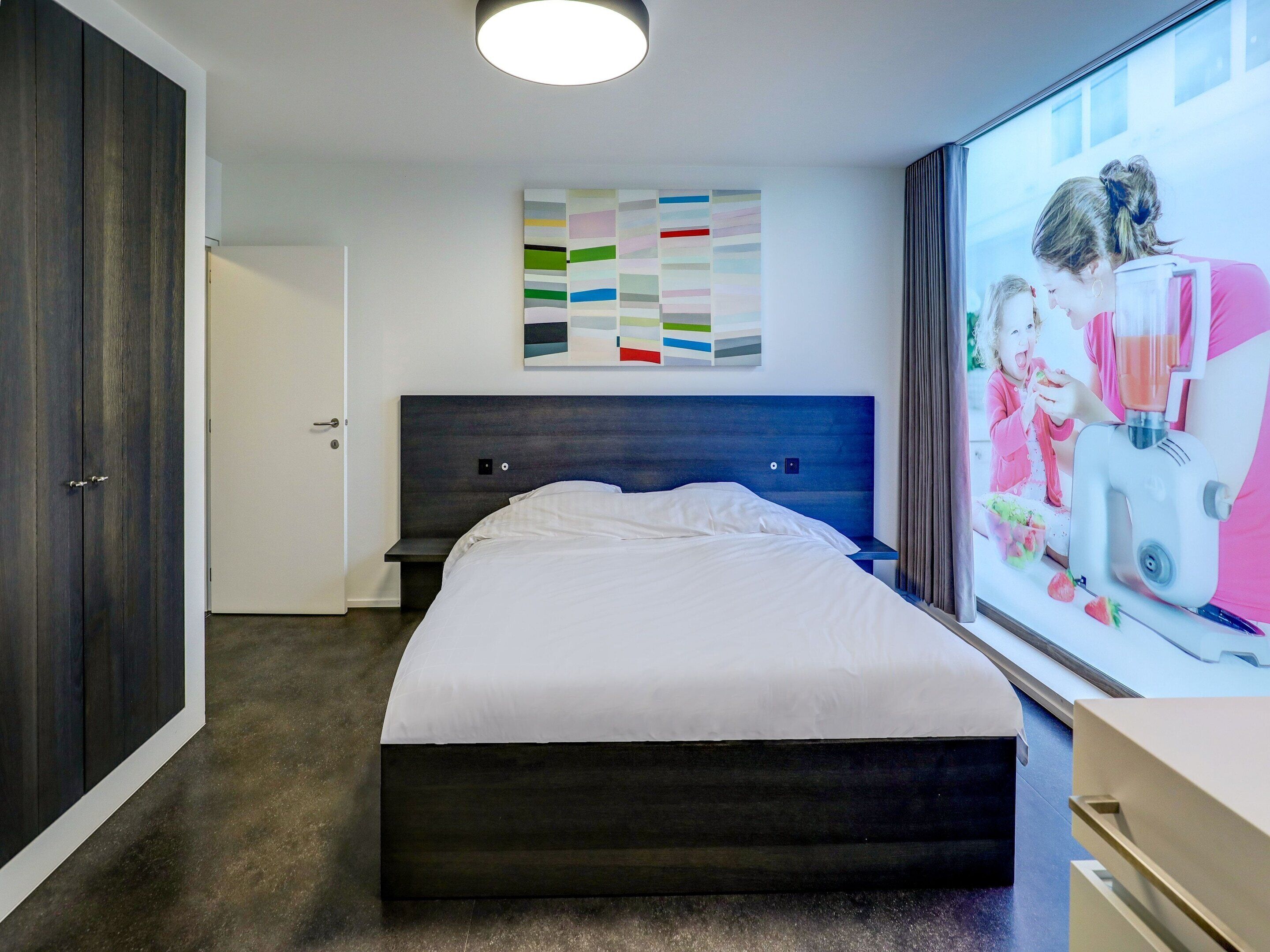Une chambre moderne avec un grand lit, une tête de lit sombre, une œuvre d’art murale et une fenêtre donnant sur une personne à l’extérieur.
