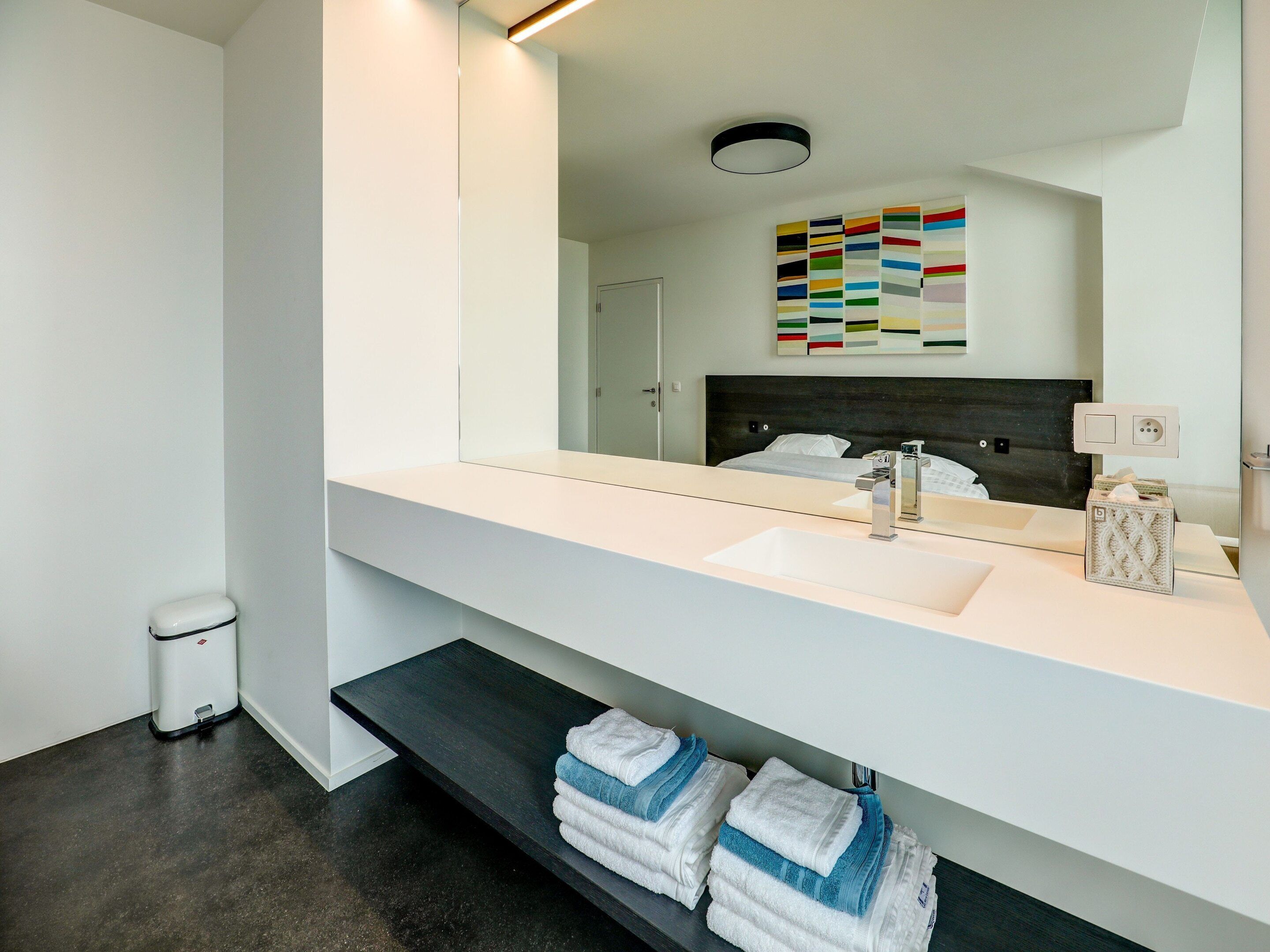 A modern bathroom with a white vanity, a large mirror, a dark headboard, and a towel rack with folded towels.