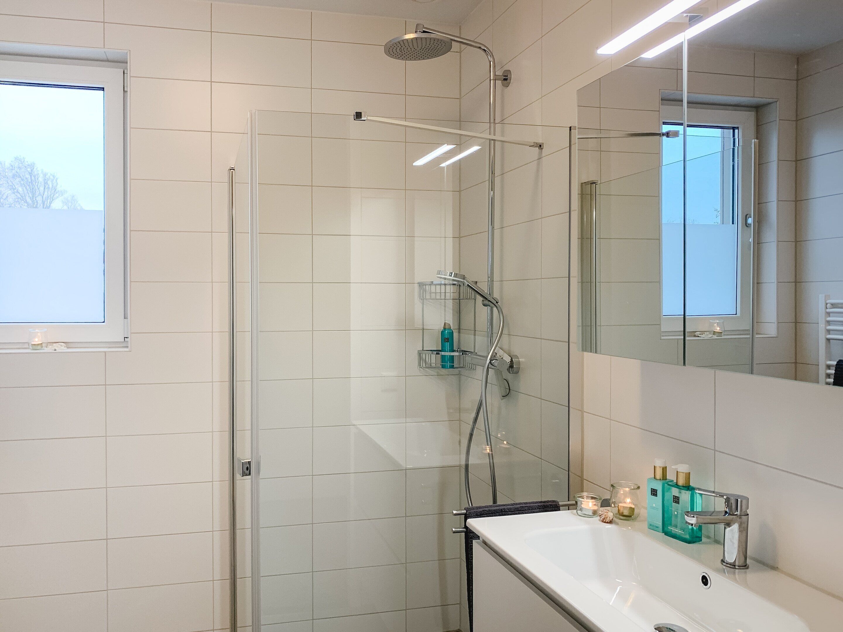 A modern bathroom with a glass-enclosed shower, a white sink with toiletries, and a large mirror.