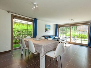 A dining area with a table set for four, surrounded by white chairs, in a room with large windows and a view of greenery outside.