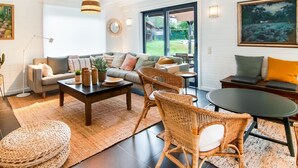 Een ruime woonkamer met een bank, salontafel, rieten stoelen en een eettafel. Er is een groot raam met uitzicht op de omgeving.