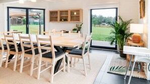 Een moderne eetruimte met een houten tafel en stoelen, een keuken met houten kasten, en uitzicht op een tuin door grote schuifdeuren.