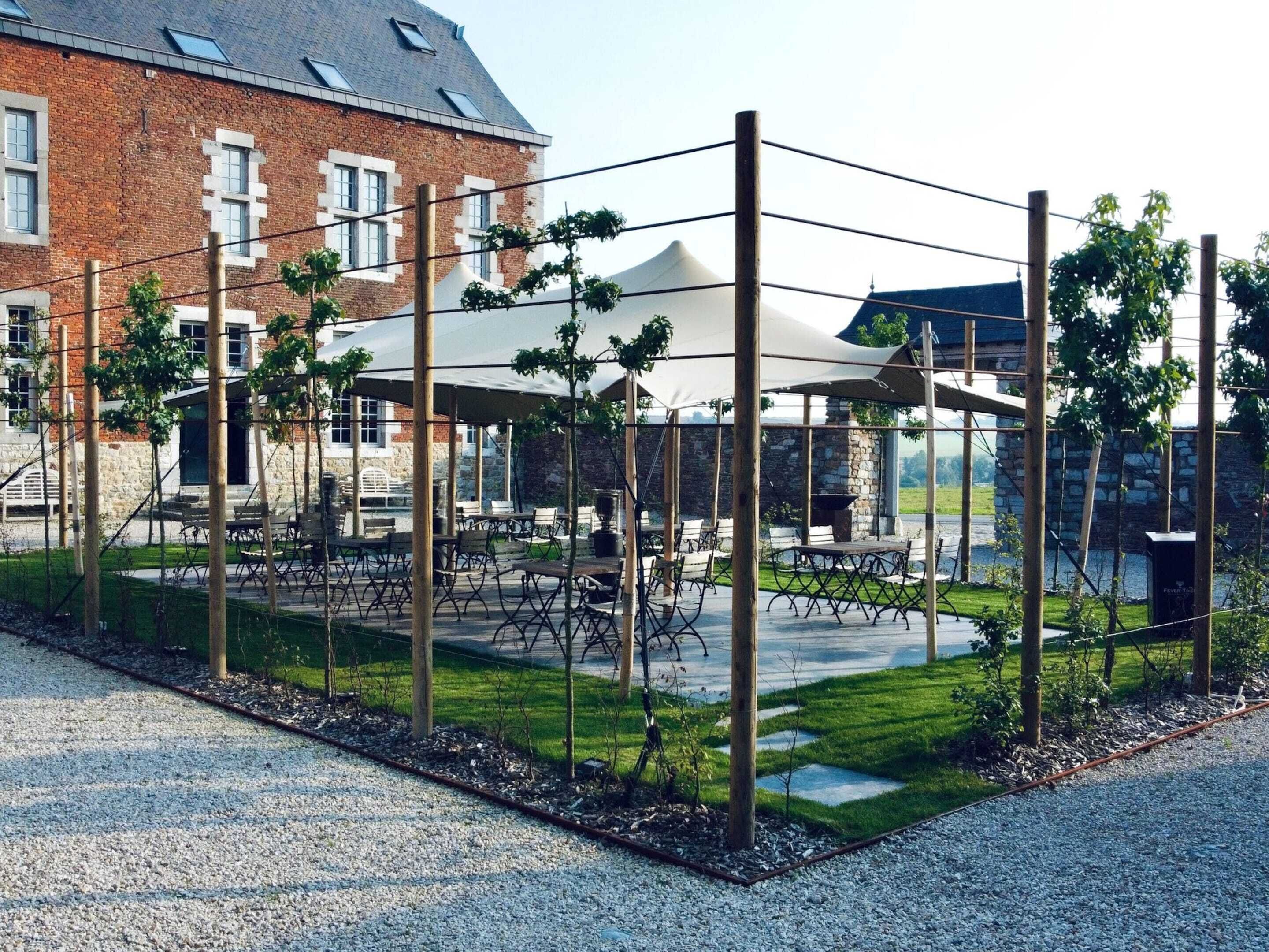 An outdoor seating area with tables and chairs under a covered structure, surrounded by a wooden fence and a brick building in the background.