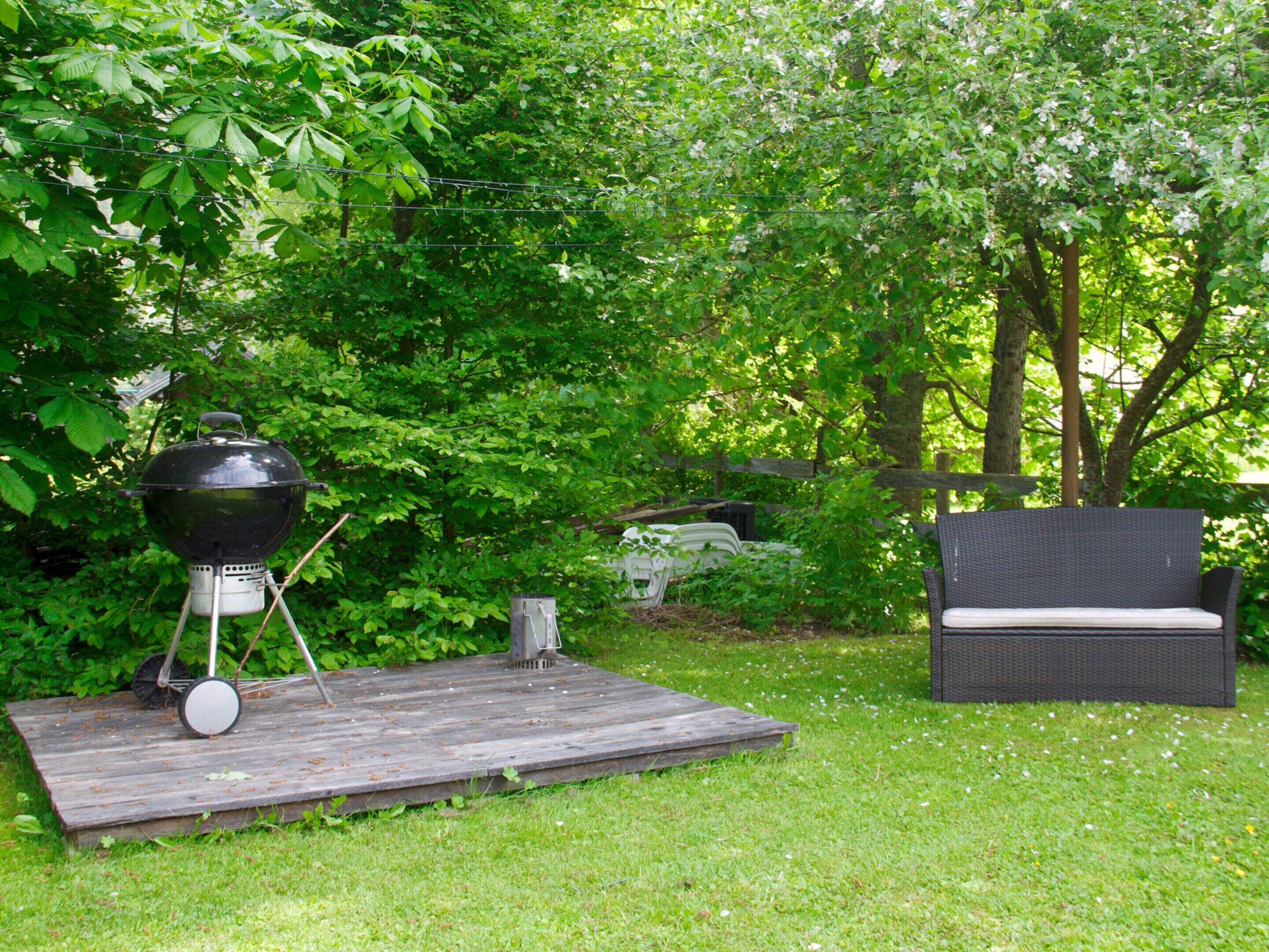 A backyard with a barbecue grill on a wooden platform, surrounded by lush greenery and a wicker chair.