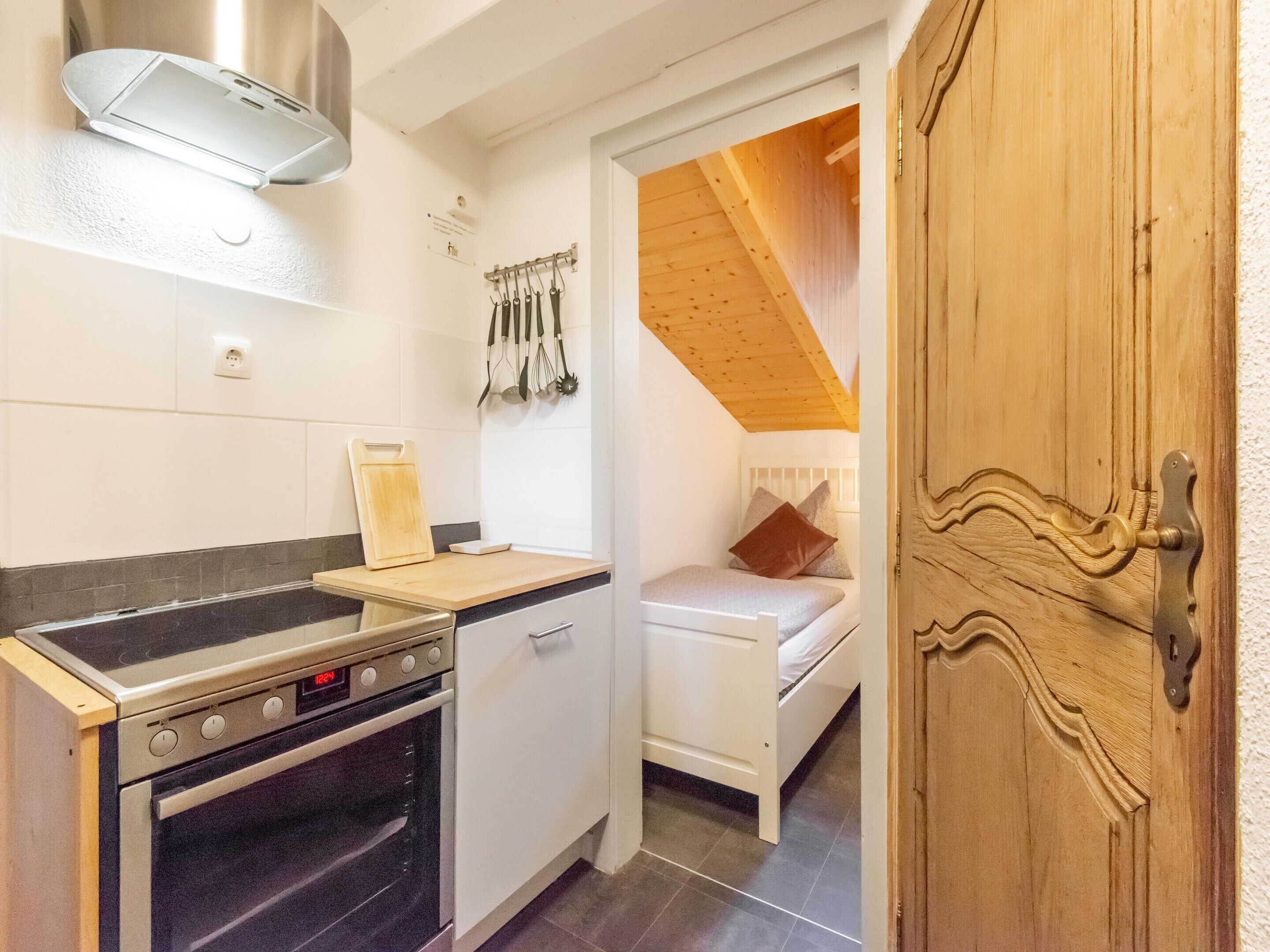 A compact kitchen with a stove, oven, and a small countertop. There is a bed with a wooden headboard in the adjacent room.