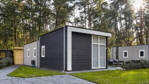 A modern, single-story cabin with a dark exterior and white trim, surrounded by a well-maintained lawn and a paved driveway. - Chalet by Forest Edge With Modern Design (Markelo)