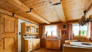 A wooden cabin interior with a kitchen area featuring wooden cabinets, a dining table, and a window with curtains. - Chalet in Jochberg Near Thurn Pass (Jochberg)