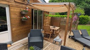 Una terraza de madera con mobiliario exterior, una pérgola con luces colgantes y un jardín delimitado por una valla de madera y plantas.
