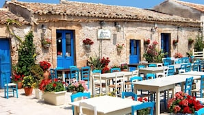 An outdoor café with white tables and blue chairs, surrounded by stone buildings with blue doors and windows. - Belvilla by OYO Agrivillage San Lorenzo (Noto)