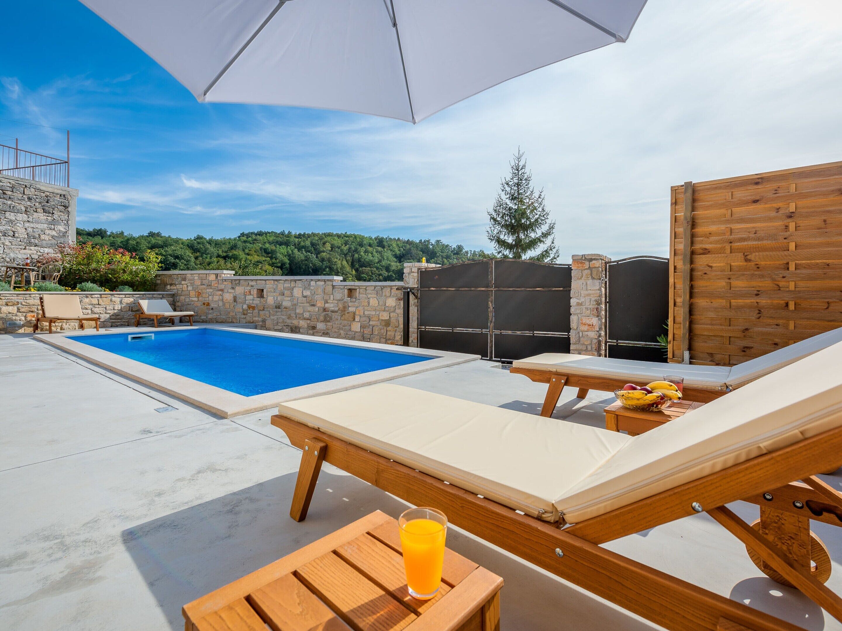 A pool area with lounge chairs and a table, a wooden fence, and a view of trees and hills.
