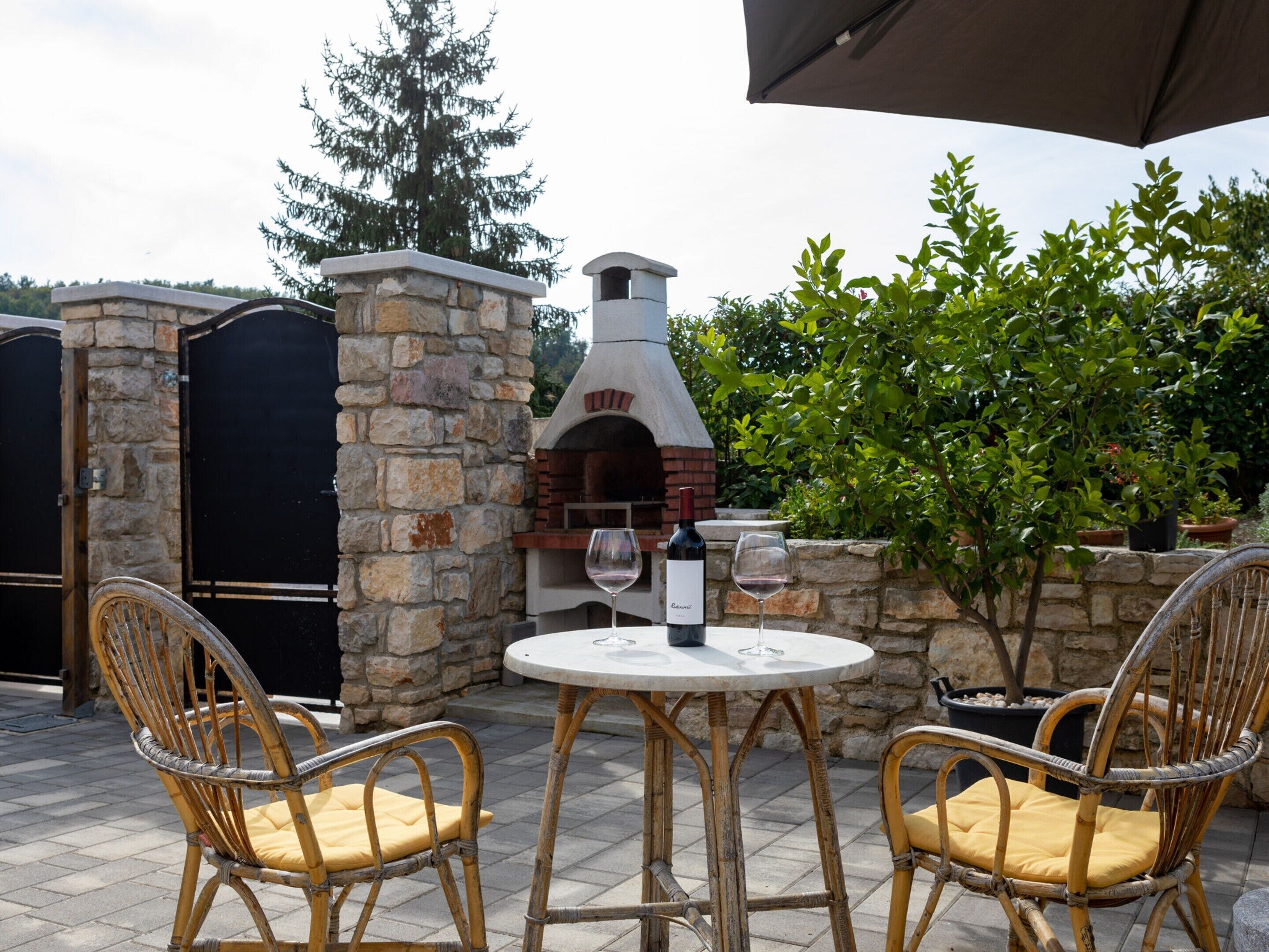 An outdoor seating area with wicker chairs and a table, a stone-built pizza oven, and a wine glass.