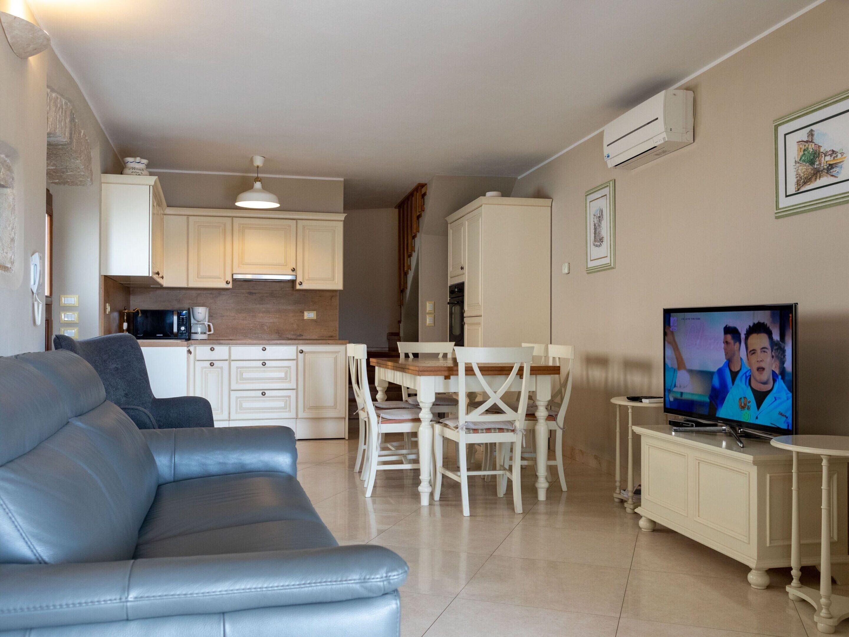 Sala de estar con sofá, mesa de comedor con sillas, cocina con armarios y electrodomésticos, y televisor sobre un mueble.