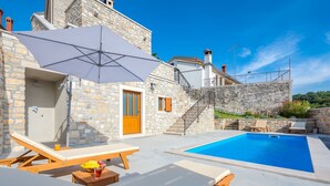 A poolside area with lounge chairs and umbrellas, stone walls, and a clear blue sky.