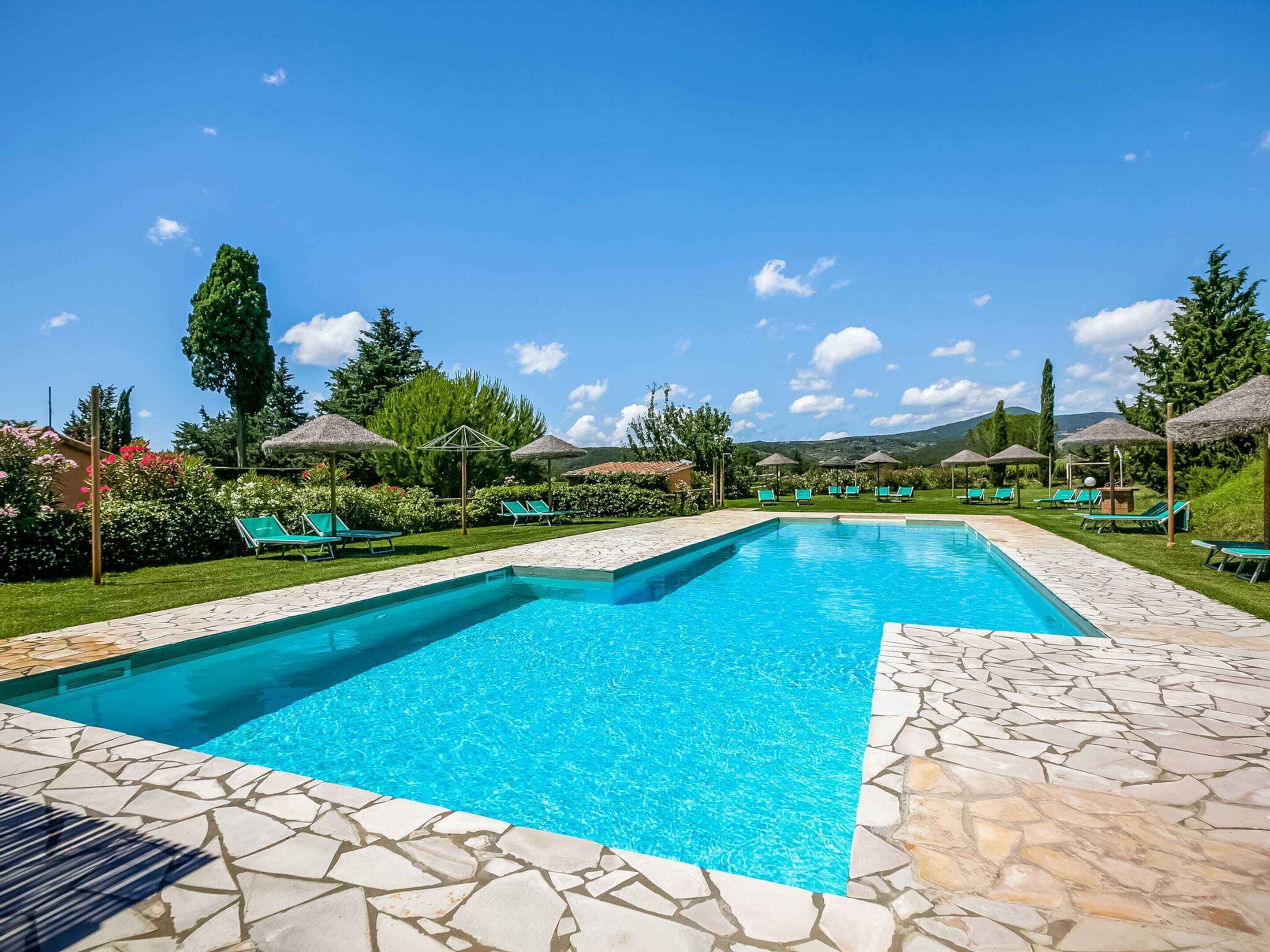 A rectangular swimming pool with a stone border, surrounded by green lounge chairs and thatched umbrellas.