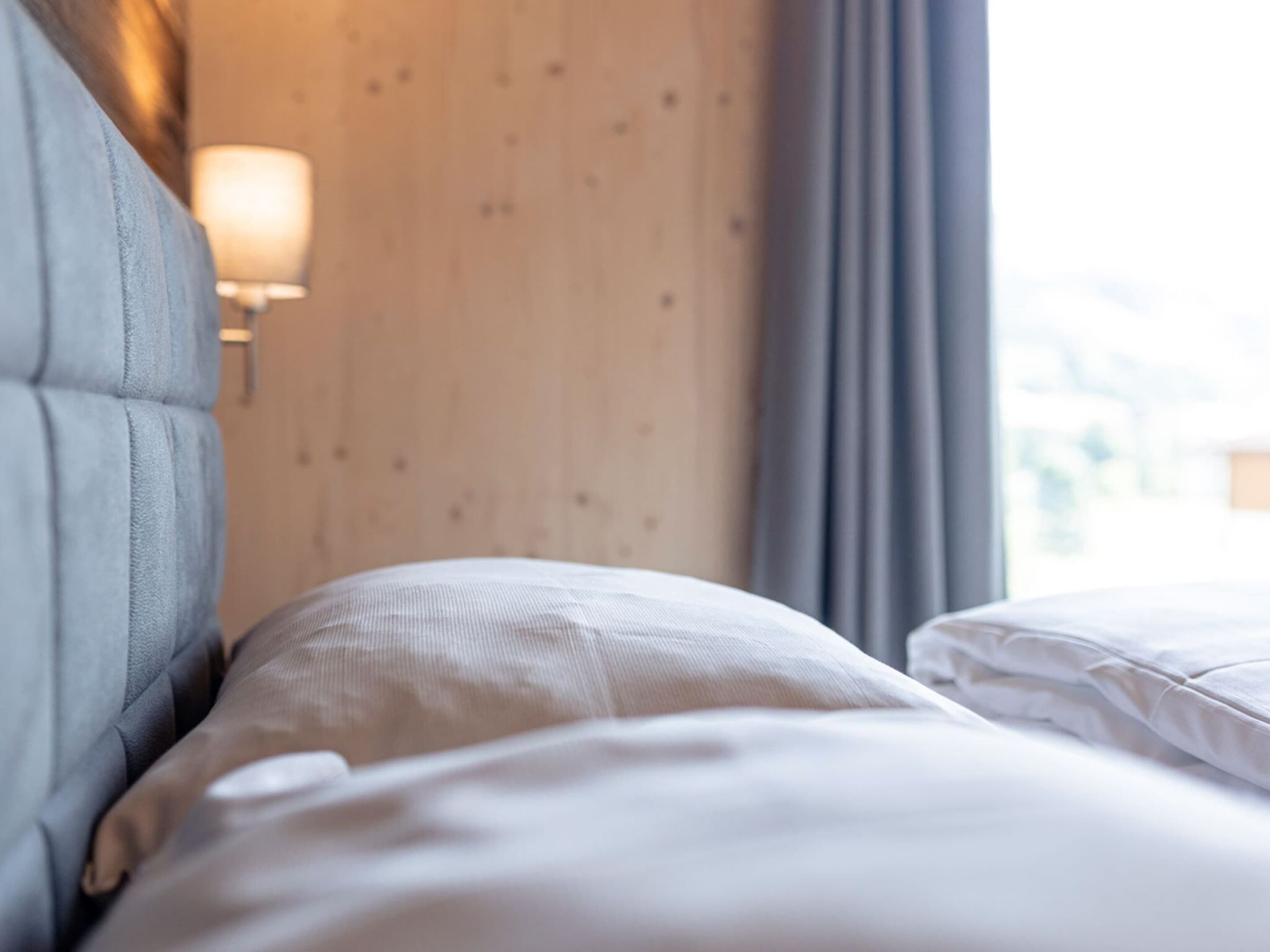 A hotel room with a neatly made bed, a lamp on the wall, and a window with curtains.
