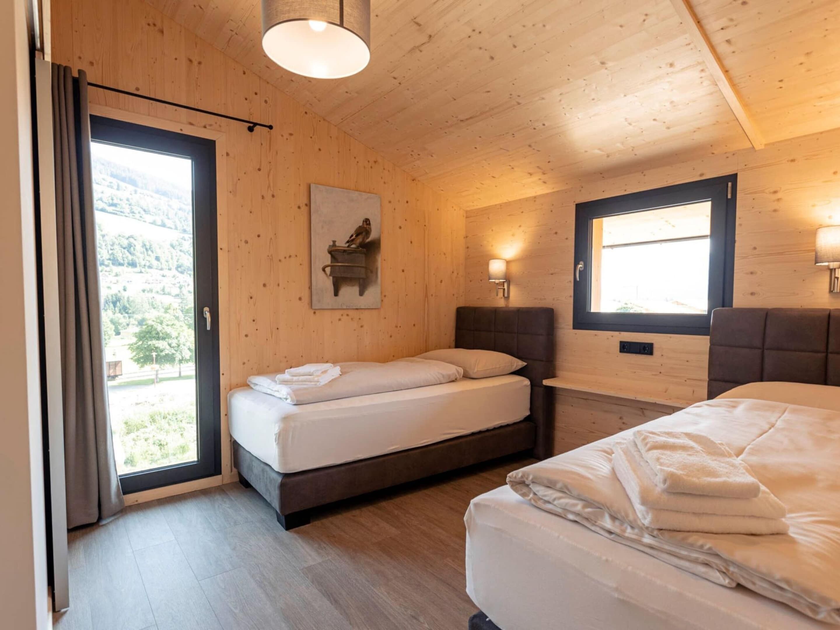 A room with two beds, wooden walls, a window with a view, and a painting on the wall.