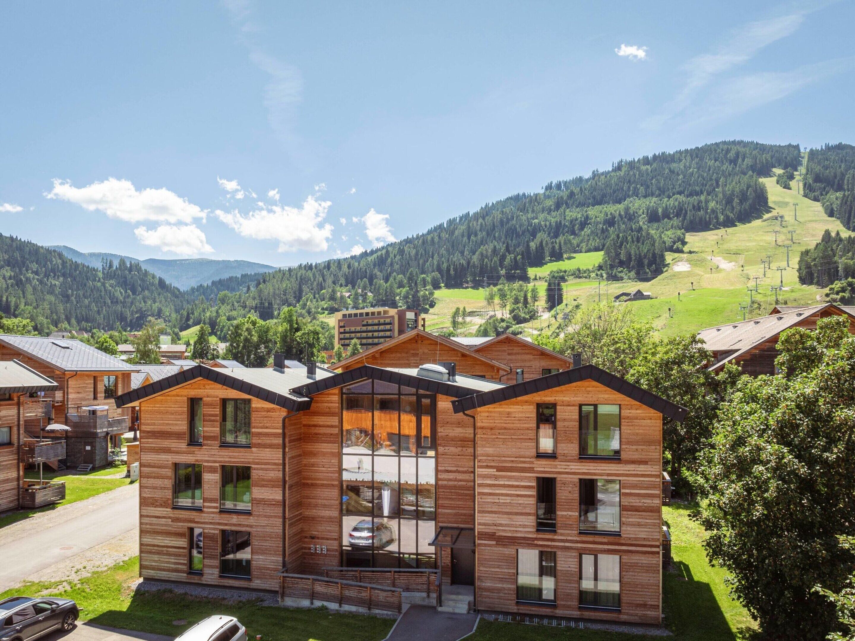 A modern wooden lodge with multiple rooms, large windows, and a view of a mountainous landscape.