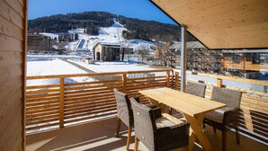 Eine Holzterrasse mit Tisch und Stühlen mit Blick auf eine verschneite Landschaft, Gebäude und eine Skipiste.