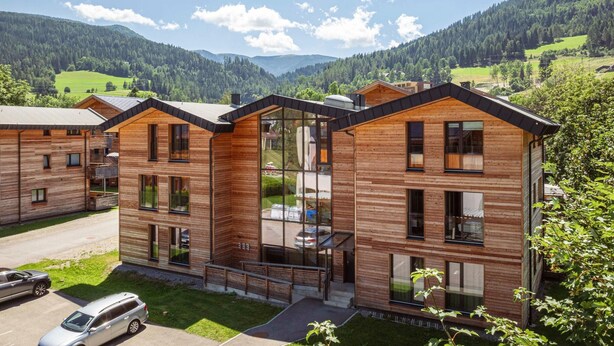 Ein modernes Holzgebäude mit großen Fenstern, einer überdachten Außenbereich und Blick auf grüne Hügel und Berge.
