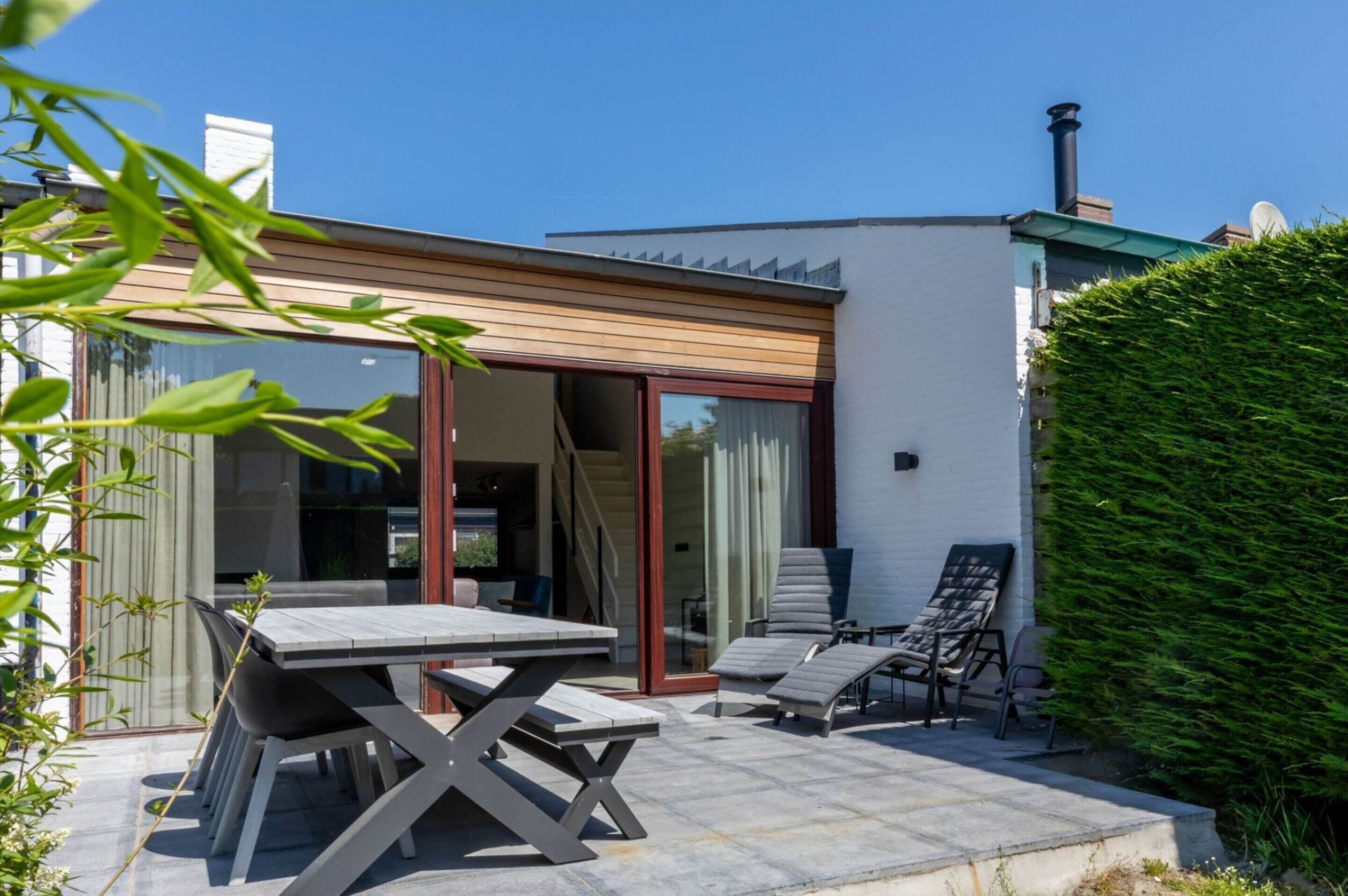 Eine moderne Außenpatio mit Esstisch und Stühlen, eine Lounge-Ecke mit Sesseln und ein großer Heckenstreifen.